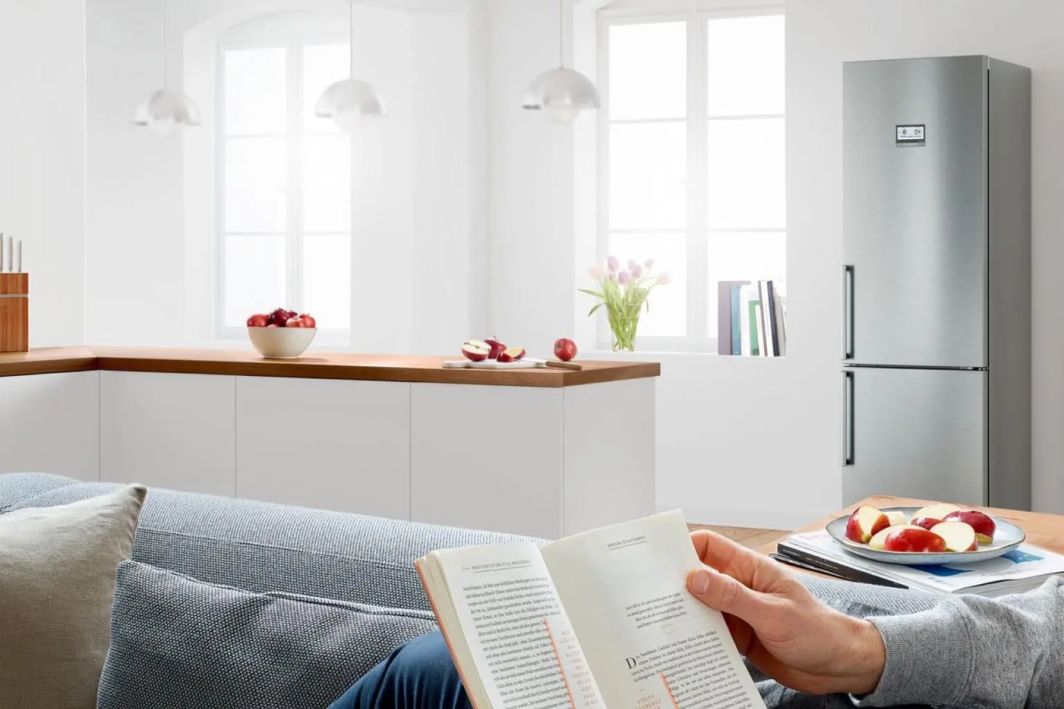 A person lies on a couch with a book and a bowl of apple slices, with a Bosch refrigerator in the background.