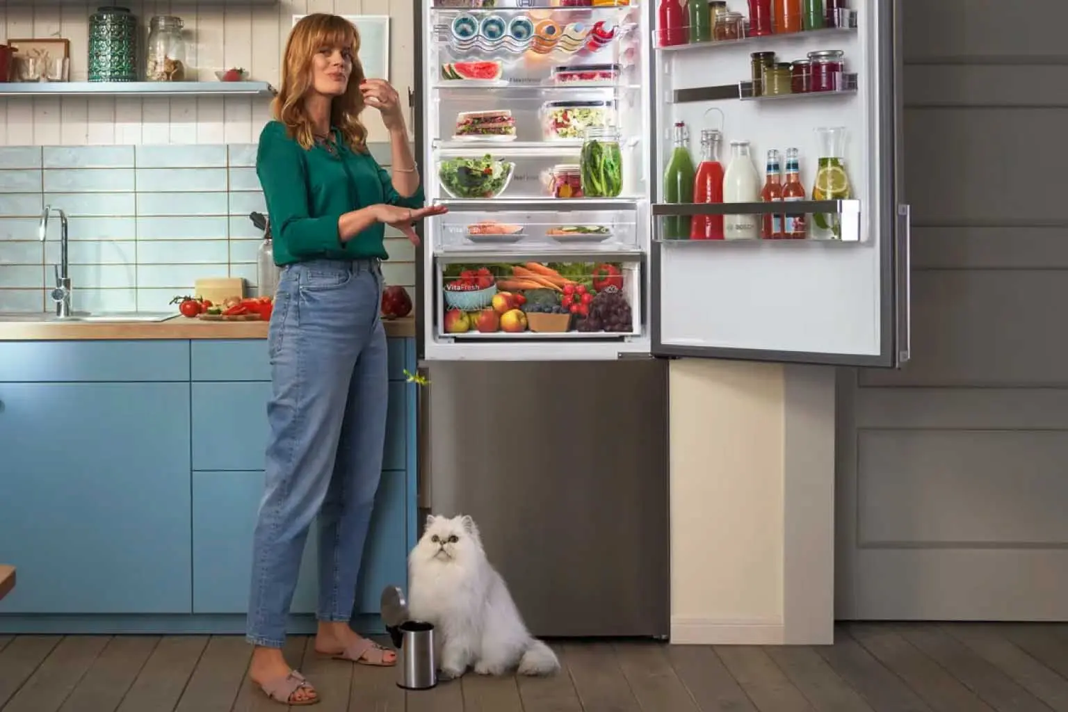 Vrouw naast open Bosch koelkast met witte kat op de grond.
