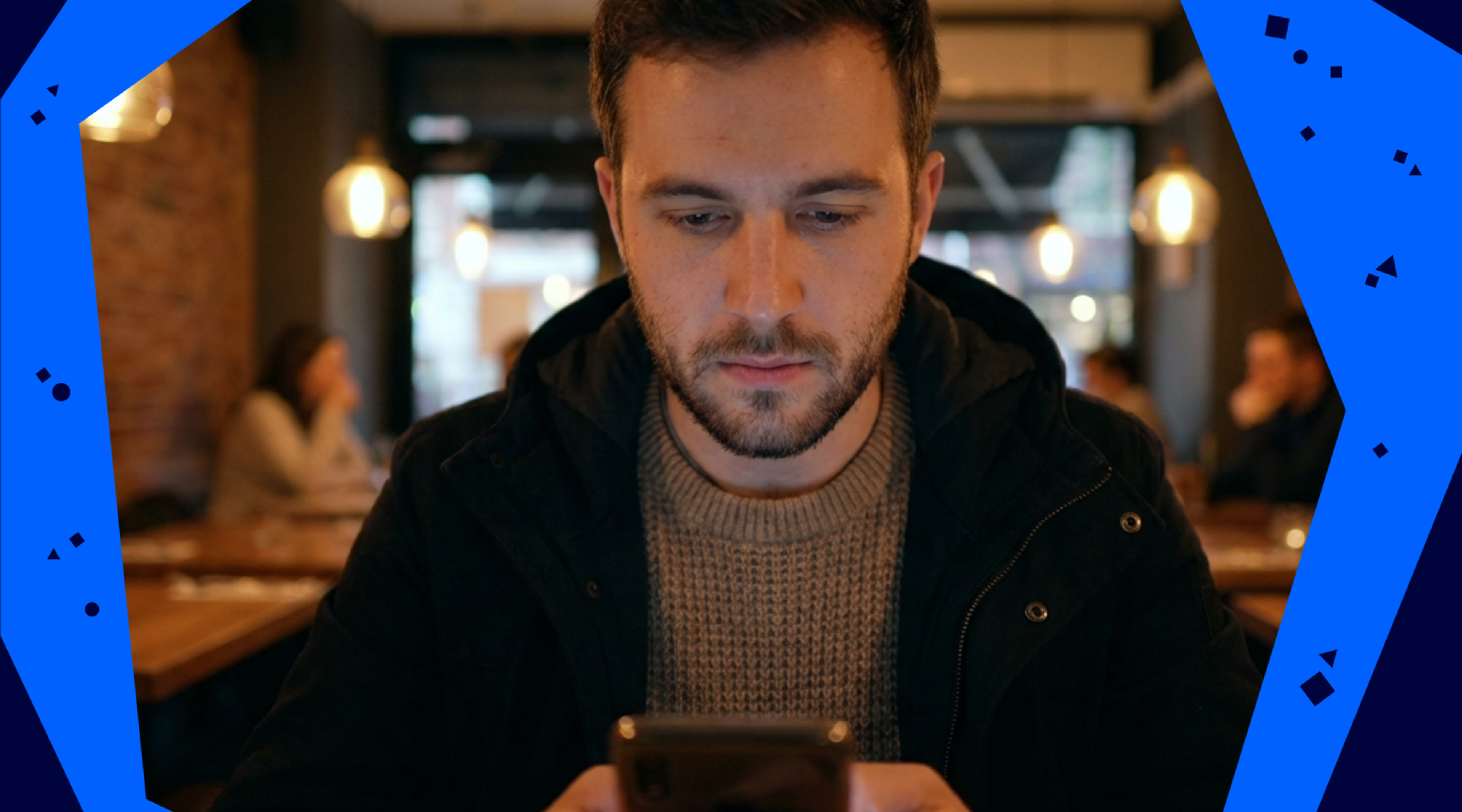 A man in a dark jacket uses a smartphone in a cozy cafe, with warm lighting and blurred background of people sitting at tables.