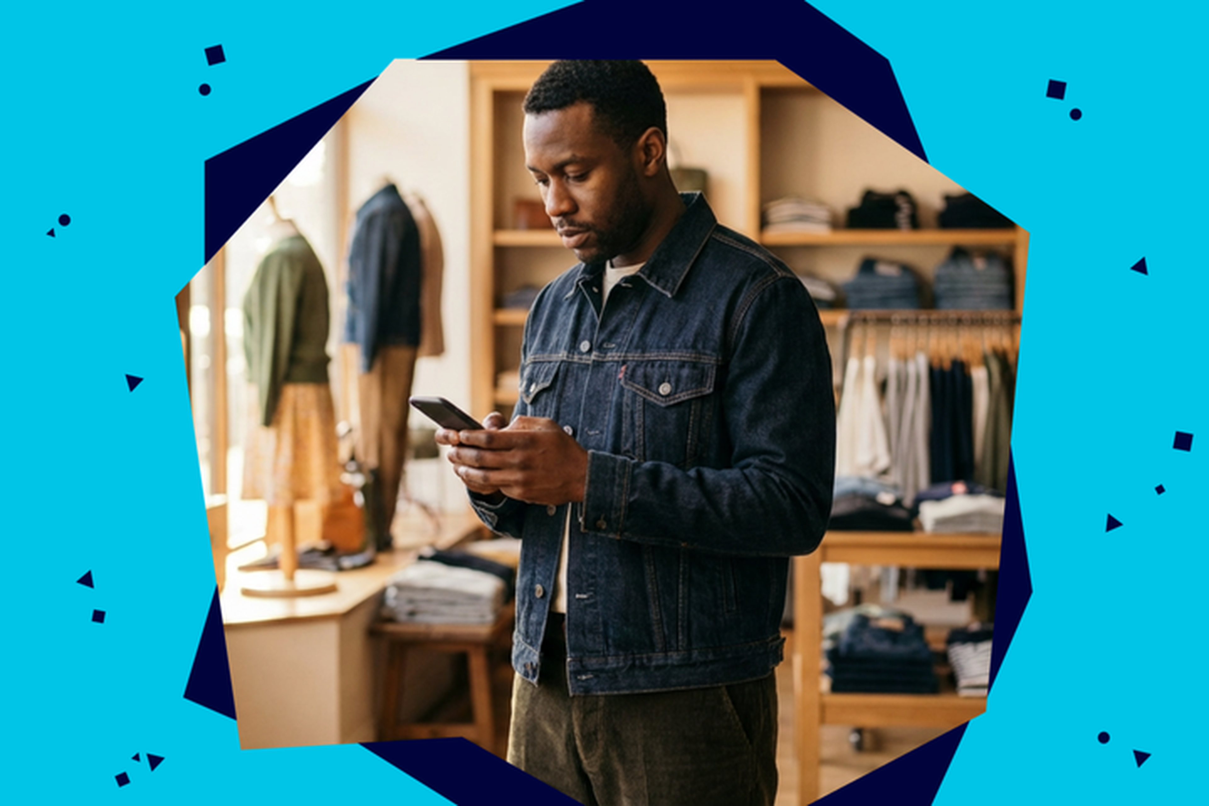Man in a denim jacket browsing his phone in a clothing store with racks of clothes in the background.