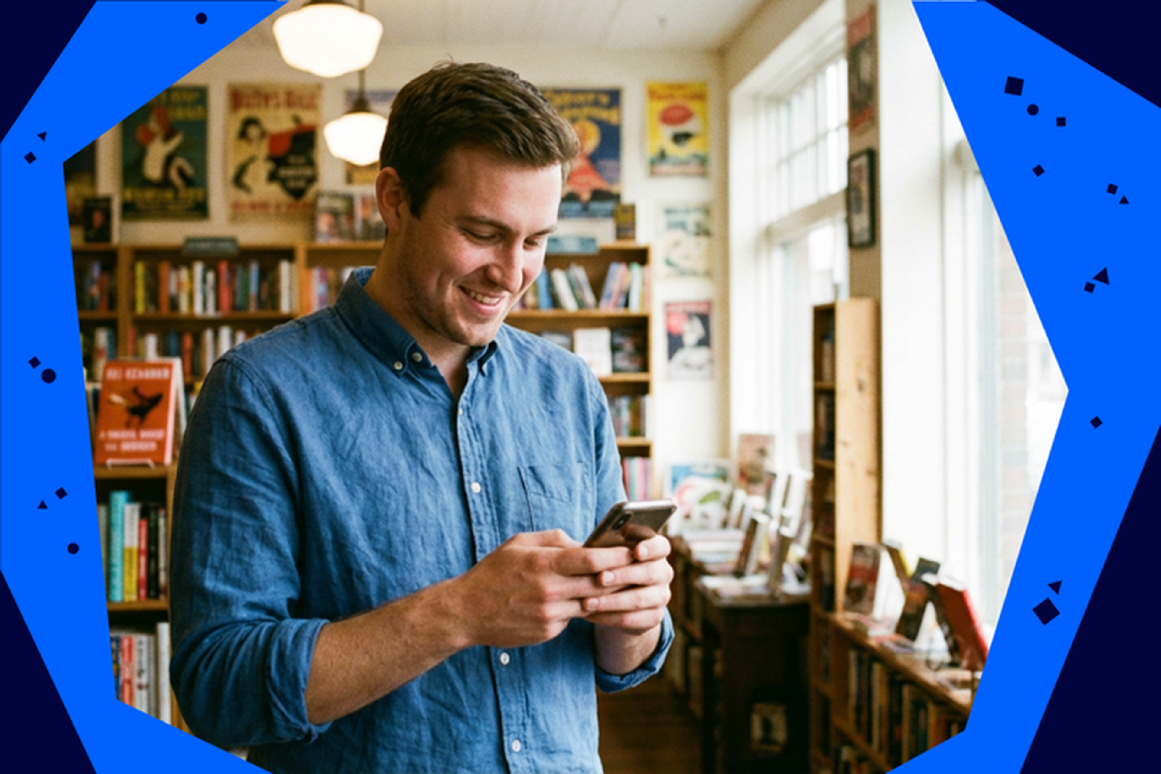 Man in a blue shirt smiling at his phone in a cozy bookstore, surrounded by posters and shelves filled with books.