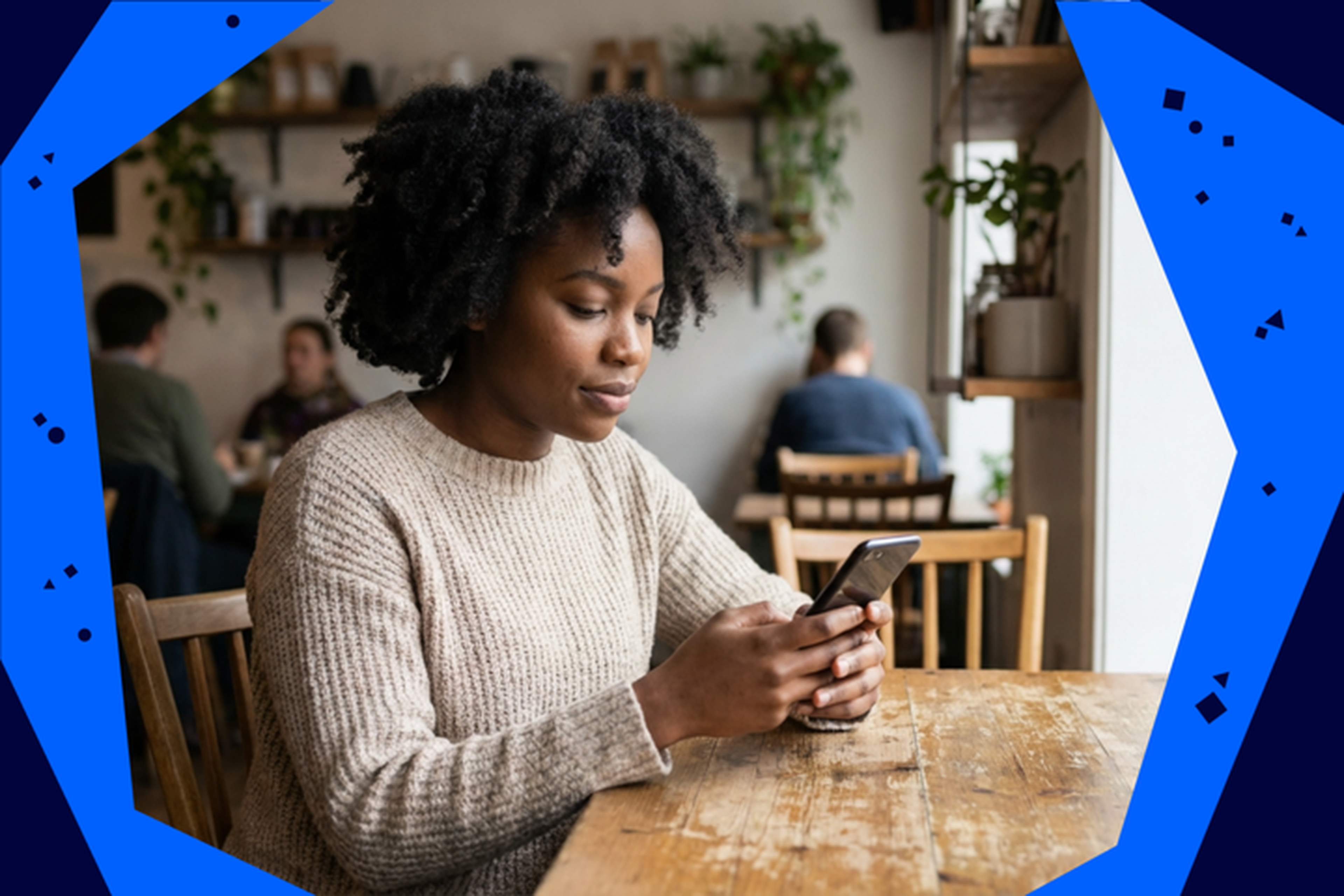 Woman with curly hair using a smartphone at a wooden table in a cozy cafe, with plants on shelves in the background.