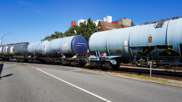 Goederentrein met cilindervormige tankwagons rijdt door een stedelijk gebied onder een heldere blauwe lucht.