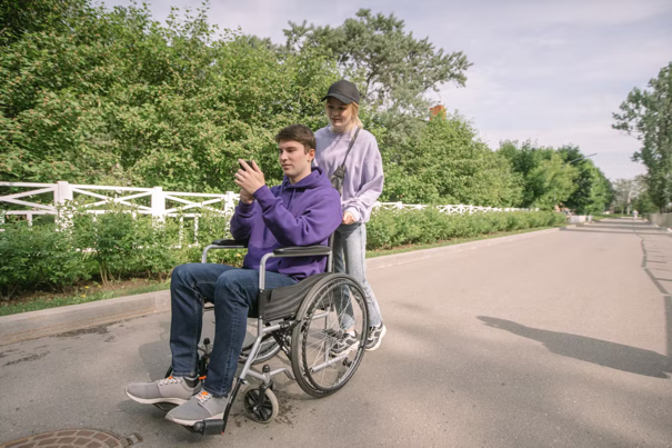 Een persoon in een rolstoel gebruikt een smartphone en wordt door iemand anders voortgeduwd in een park, omringd door groen en een wit hek.