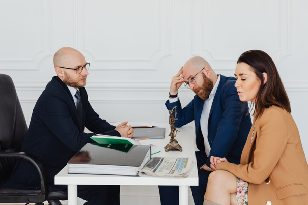 Drie mensen zitten in een serieus overleg aan een tafel met documenten en een krant, in een professionele kantooromgeving in een advocatenkantoor.