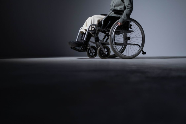 Persoon in een rolstoel op een schemerig verlichte vloer, met een donker jasje en lichte broek, tegen een minimalistische achtergrond.