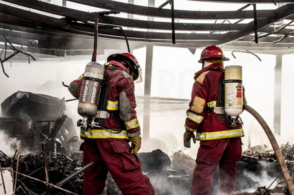 Twee brandweerlieden in rode uitrusting en met helmen, voorzien van ademluchtflessen, staan tussen puin en rook onder een beschadigde metalen constructie.