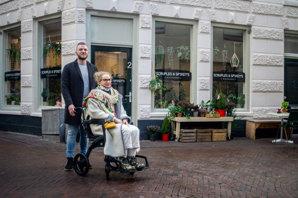 Een man staat achter een vrouw in een rolstoel buiten een plantenwinkel in een straat met bakstenen, omringd door potplanten.
