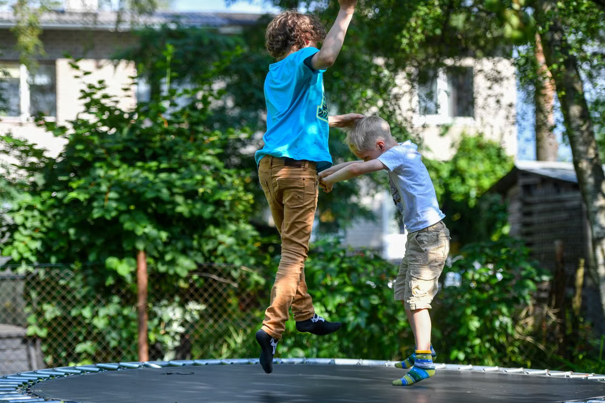 Twee kinderen springen op een trampoline in een achtertuin, met bomen en een hek op de achtergrond.