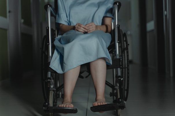 Persoon in een ziekenhuisjas zit in een rolstoel, met de handen gevouwen en de voeten op de voetsteunen, in een schemerig verlichte gang.