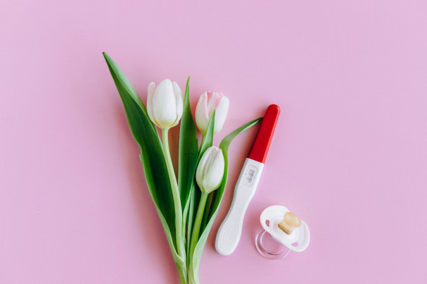 Drie witte tulpen, een positieve zwangerschapstest en een fopspeen op een roze achtergrond.