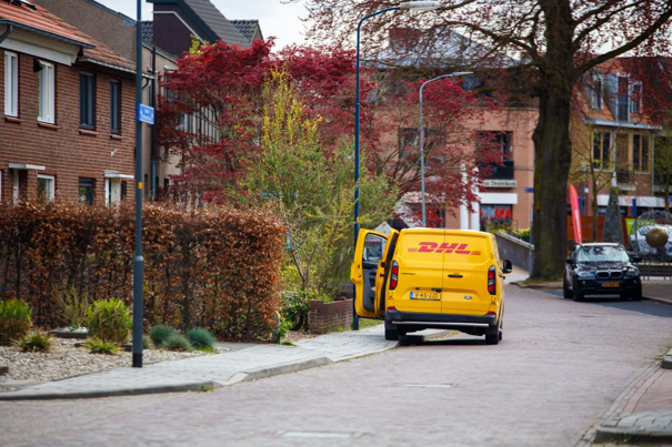 Gele DHL-bestelwagen geparkeerd in een woonstraat met open deuren. In de buurt staat een boom met rode bladeren.