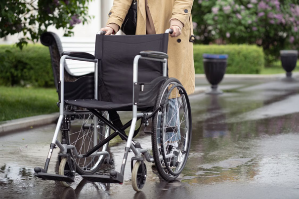 Een persoon in een beige jas duwt een lege rolstoel over een nat pad in een park, met groen en bankjes op de achtergrond.