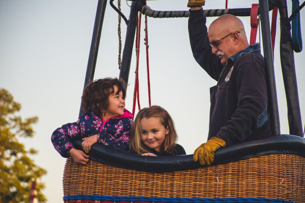 Twee glimlachende kinderen en een volwassene in een rieten mand van een heteluchtballon, met bomen zichtbaar op de achtergrond.