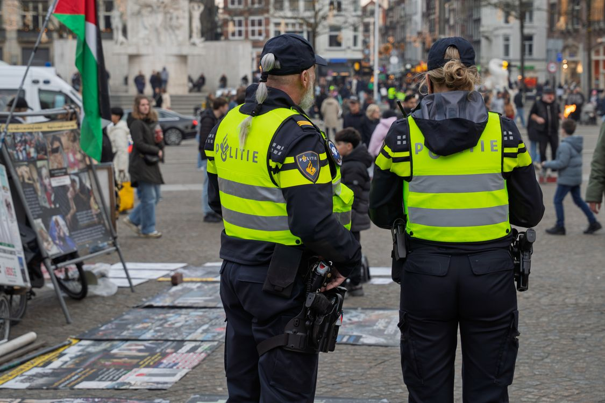 Twee politieagenten in fluohesjes observeren een openbaar plein met posters en mensen in de buurt, inclusief een Palestijnse vlag.