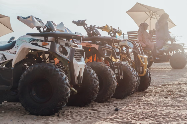Quads die op een rij geparkeerd staan op zanderig terrein, met op de achtergrond twee mensen op een bankje onder parasols.