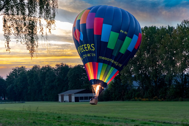 Een kleurrijke heteluchtballon landt op een grasveld bij zonsondergang, met bomen en een klein gebouw op de achtergrond.