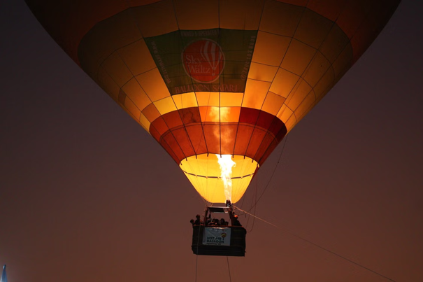 Een kleurrijke heteluchtballon met een brandende brander stijgt op tegen een schemerige hemel, met meerdere mensen in de mand eronder.