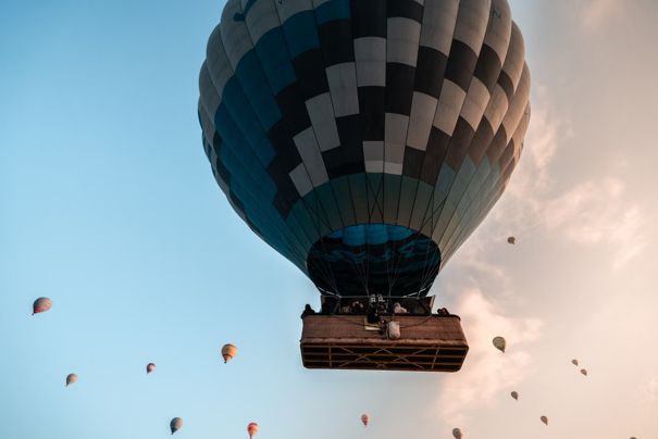 Grote heteluchtballon met een zwart-turkoois patroon in de lucht, omringd door kleinere kleurrijke ballonnen bij zonsopgang of zonsondergang.