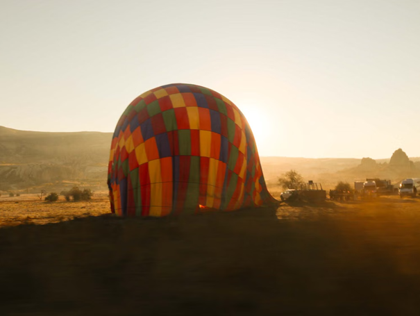 Een leeggelopen, kleurrijke heteluchtballon ligt op een veld bij zonsopgang, met voertuigen in de buurt en een heuvelachtig landschap op de achtergrond.