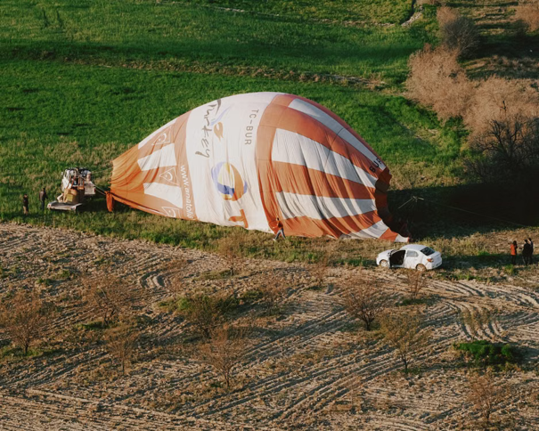 Een leeggelopen heteluchtballon ligt op het gras, met enkele mensen en een witte auto in de buurt, omringd door velden en verspreide bomen.