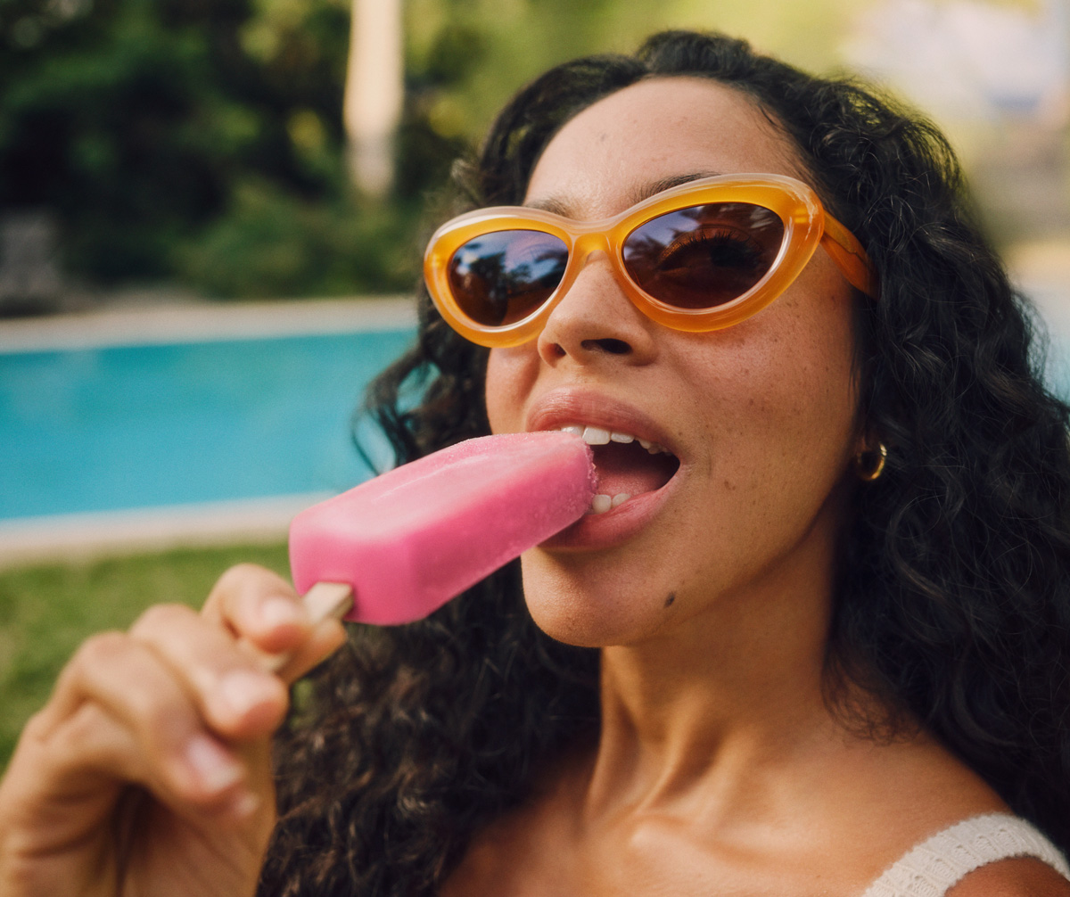 Eine Frau mit lockigen Haaren trägt orangefarbene Sonnenbrillen und genießt an einem sonnigen Tag am Pool ein pinkes Eis am Stiel.