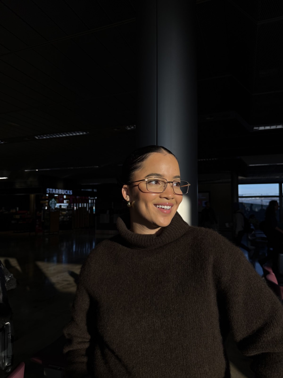 Donna sorridente con occhiali e un maglione marrone scuro è in una zona soleggiata, con un bar e delle persone sullo sfondo.