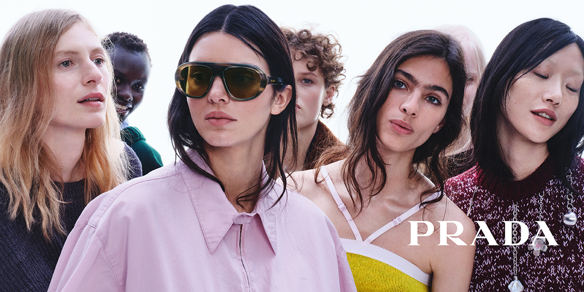 Eine Gruppe bunt und modisch gekleideter Personen posiert gemeinsam. Eine Person trägt eine große Sonnenbrille. In der unteren rechten Ecke ist „PRADA“ zu sehen.