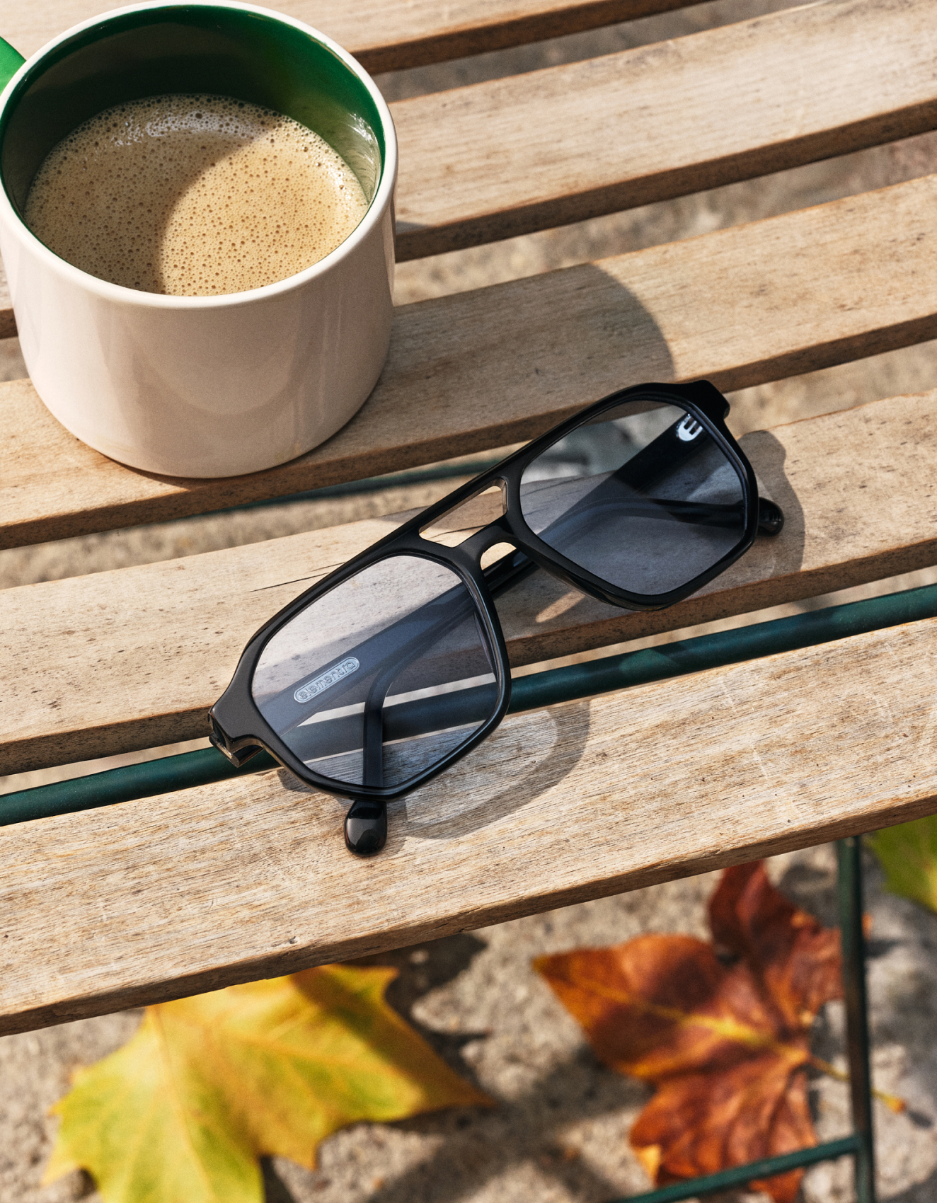 Eine Sonnenbrille und eine Kaffeetasse stehen auf einer Holzbank, während Herbstblätter auf dem Boden darunter verstreut liegen.