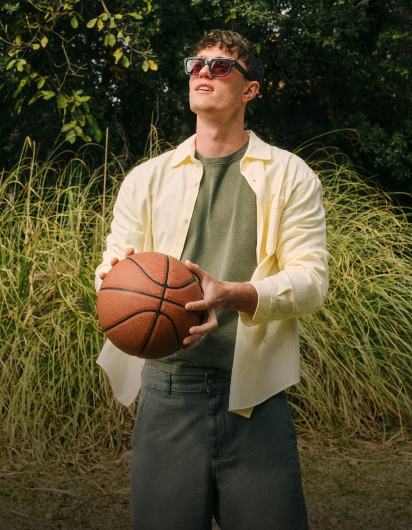 Eine Person mit Sonnenbrille hält einen Basketball und trägt ein gelbes Hemd über einem grünen T-Shirt. Er steht im Freien vor einem grünen, bewachsenen Hintergrund.