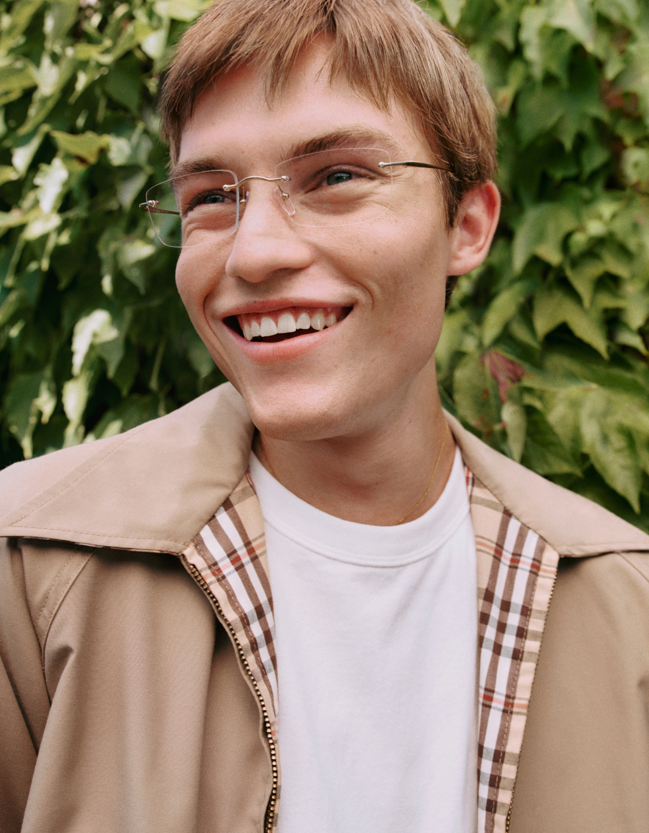 Eine lächelnde junge Person mit Brille und kurzen Haaren steht vor einer üppigen, grünen, mit Efeu bewachsenen Wand und trägt eine beige Jacke über einem weißen Hemd.
