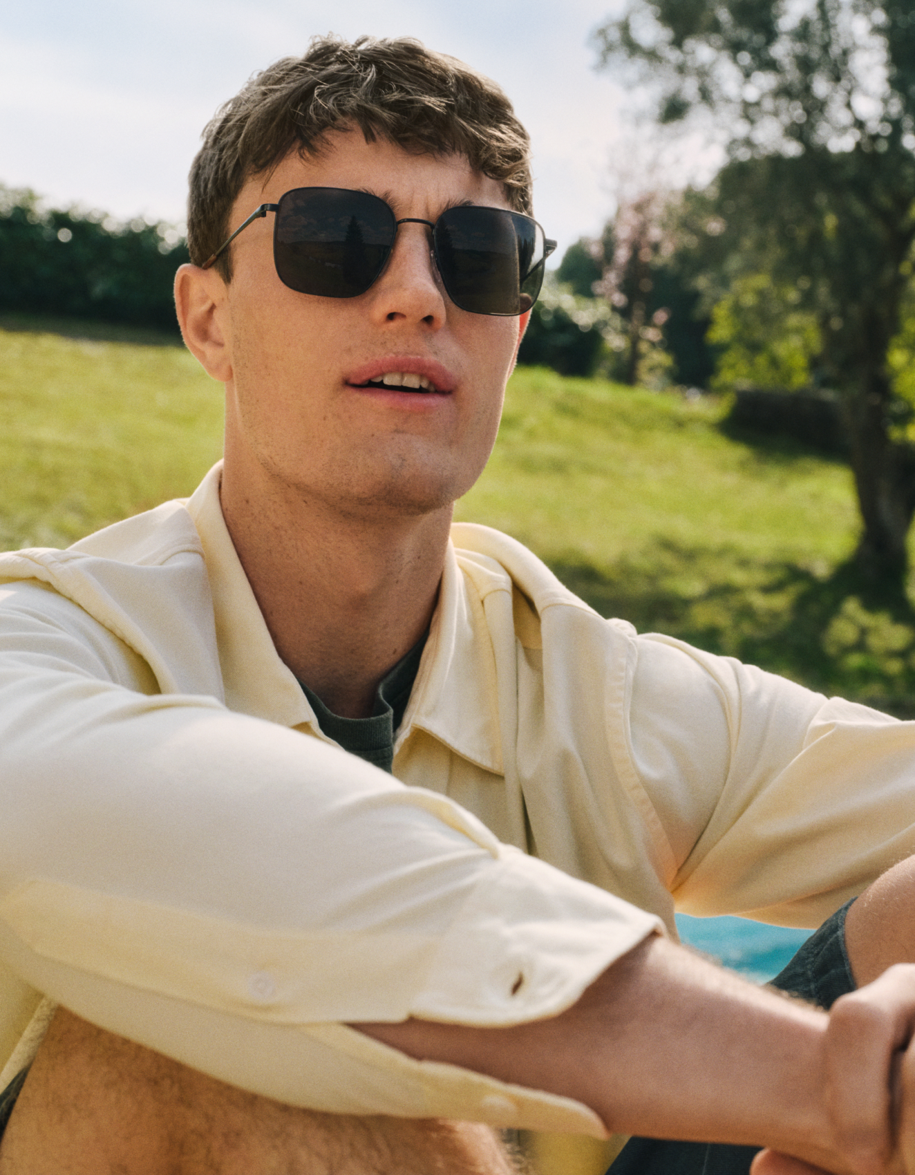 Un giovane con occhiali da sole e una camicia giallo chiaro è seduto all'aperto in una giornata soleggiata; sullo sfondo si vedono erba e alberi.