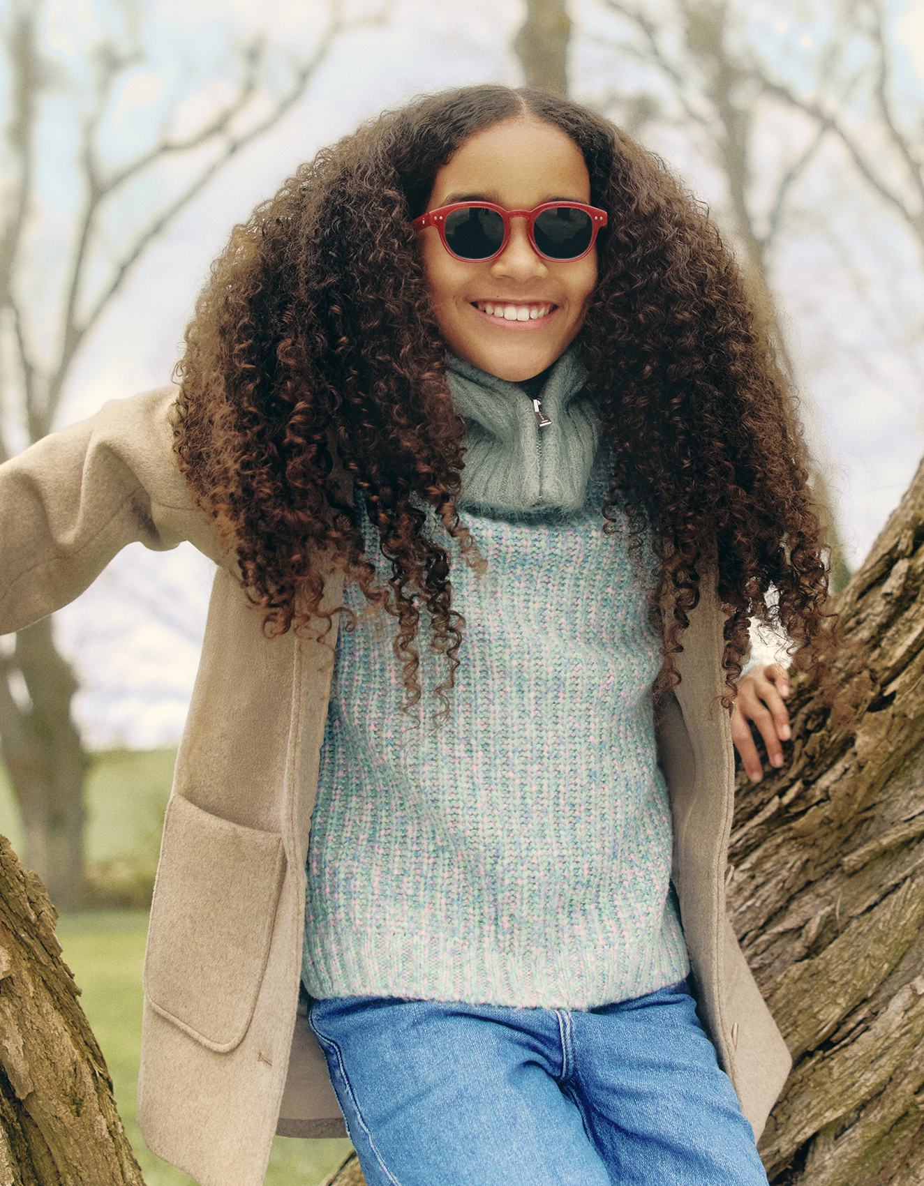 Ein lächelndes Mädchen mit langen Locken und roter Sonnenbrille lehnt draußen an einem Baum und trägt einen beigen Mantel, einen blauen Pullover und Jeans.