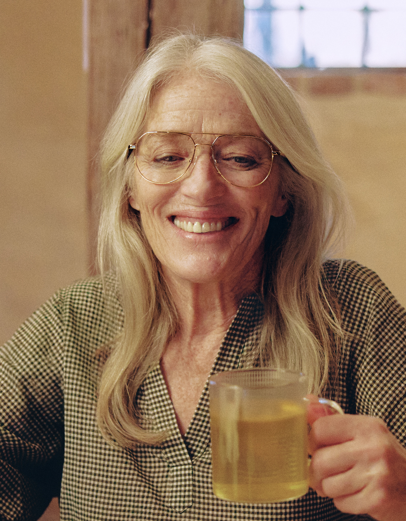 Dame mit langem grauen Haar und Brille, lächelt und hält eine Tasse Tee.