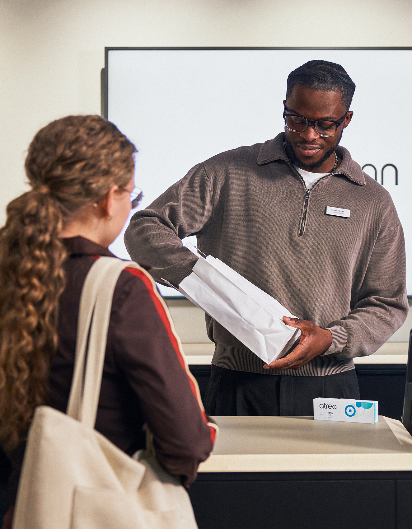 Ein Ladenangestellter mit Brille und Namensschild reicht einer Person mit langen Haaren eine Papiertüte.