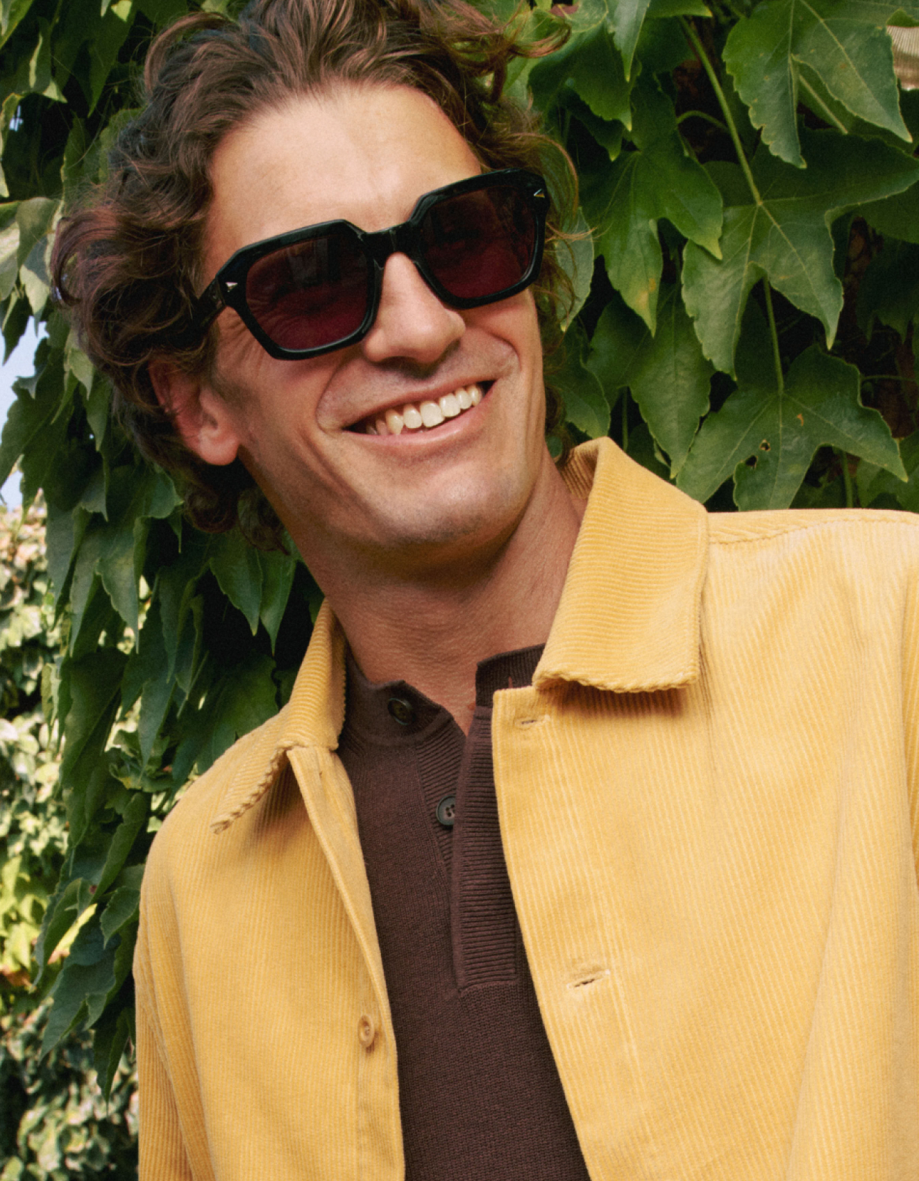 Ein lächelnder Mann mit gewelltem Haar, der eine Sonnenbrille, eine gelbe Jacke und ein braunes Hemd trägt und vor grünen Blättern steht.