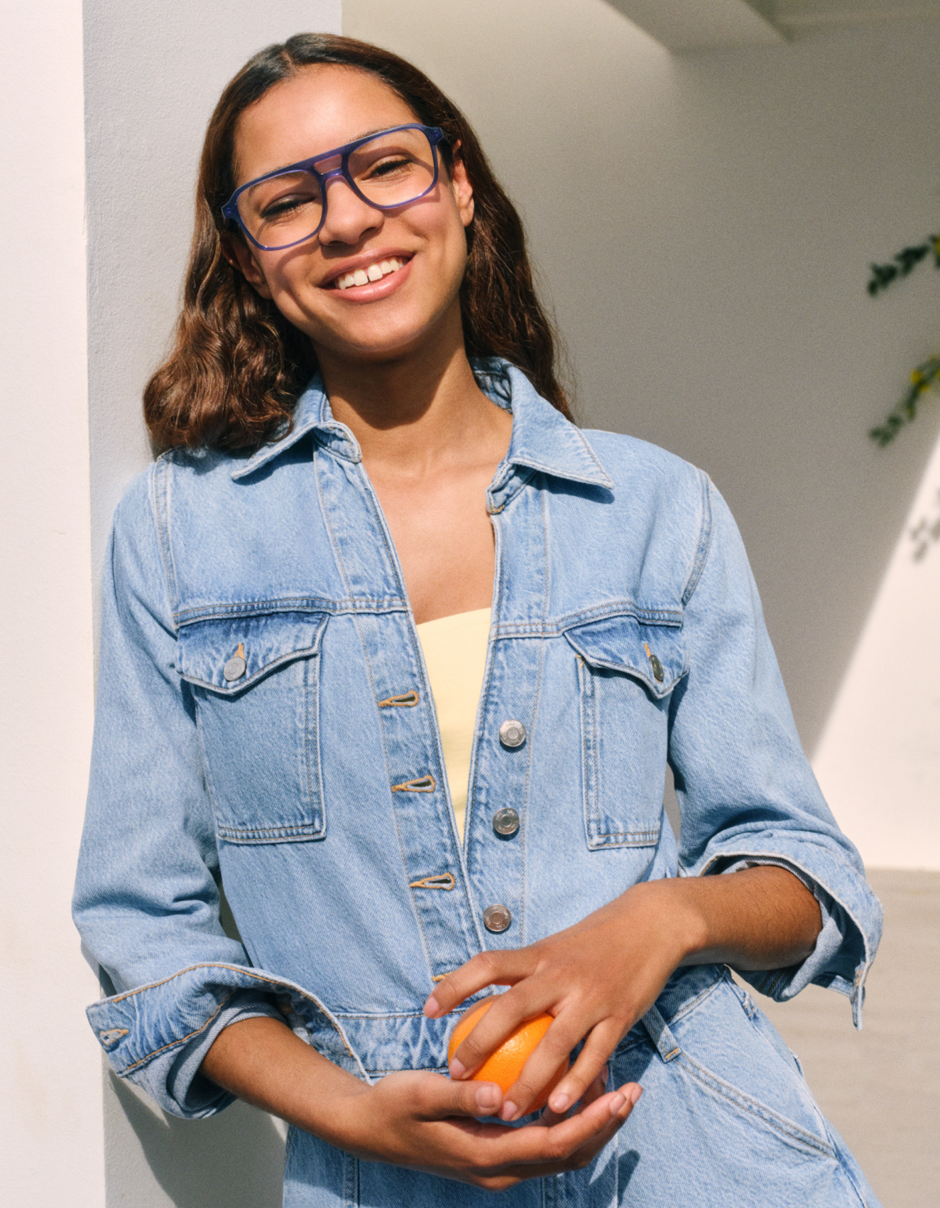Eine Frau mit einer Jeansjacke und einer Brille lächelt, während sie eine Orange in der Hand hält. Sie steht vor einer von Sonnenlicht beleuchteten weißen Wand.