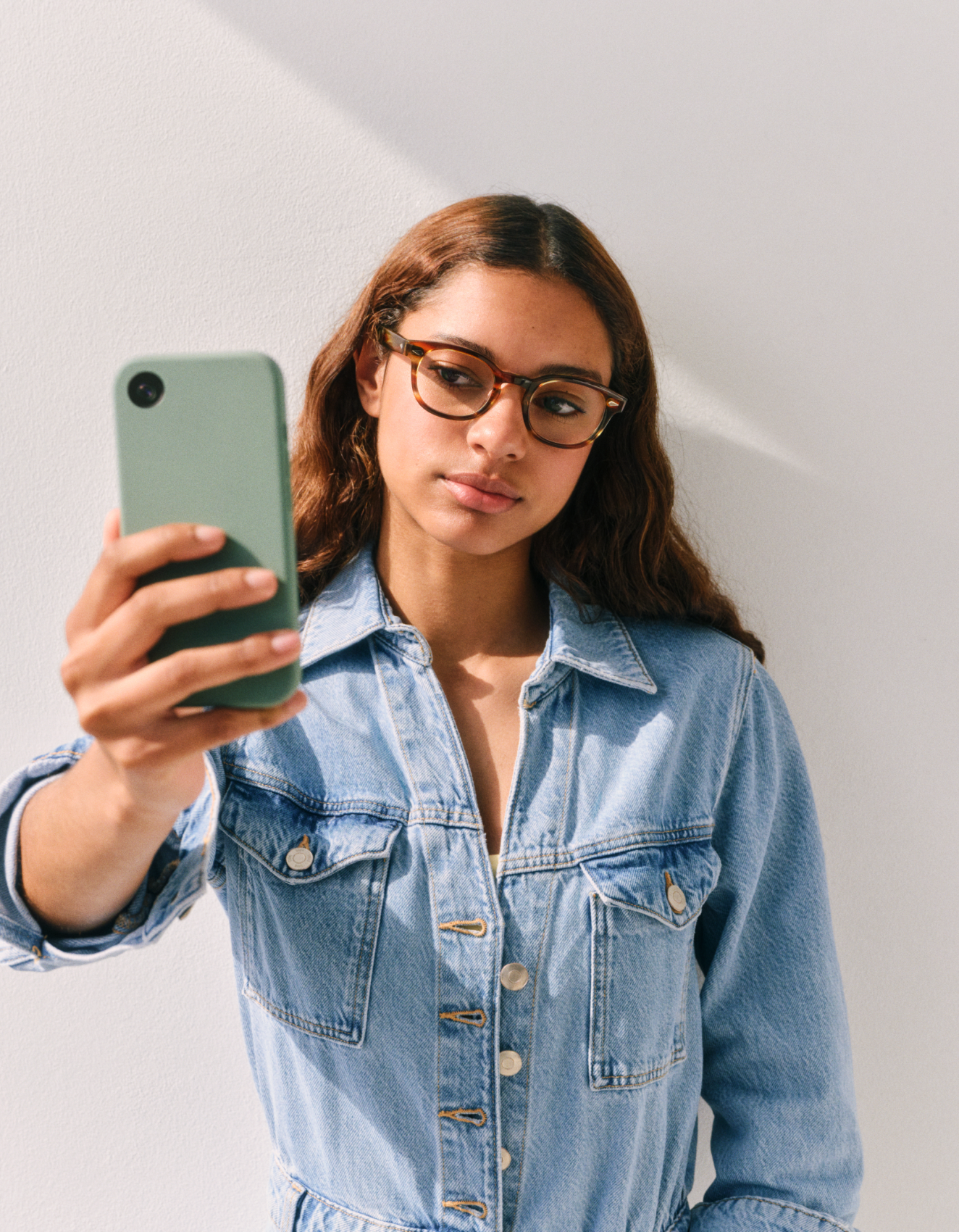 Osoba w okularach i dżinsowej kurtce robi sobie selfie na tle jasnej ściany, trzymając w dłoni smartfon w zielonym etui.