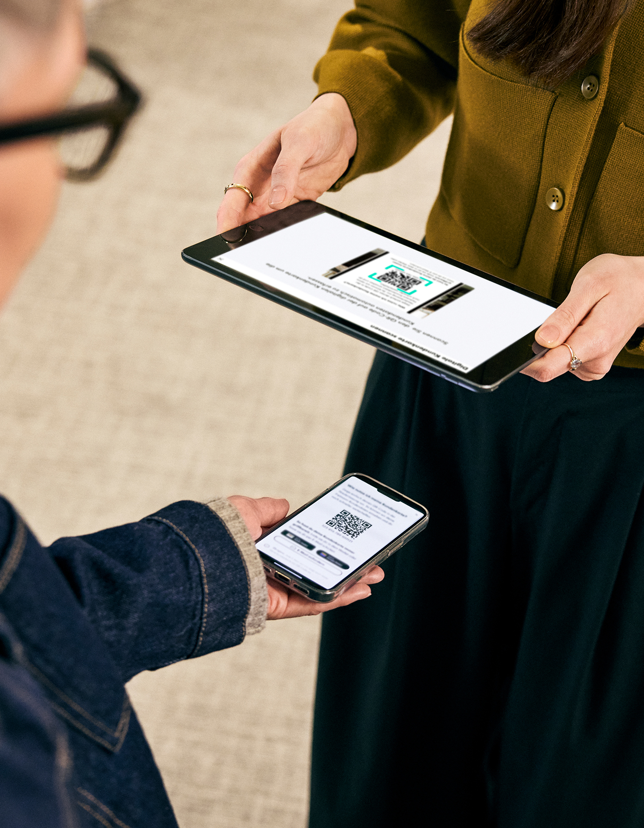 Zwei Personen tauschen Informationen über ein Tablet und ein Smartphone aus. Auf dem Tablet wird eine Website angezeigt, während auf dem Smartphone ein QR-Code zu sehen ist.