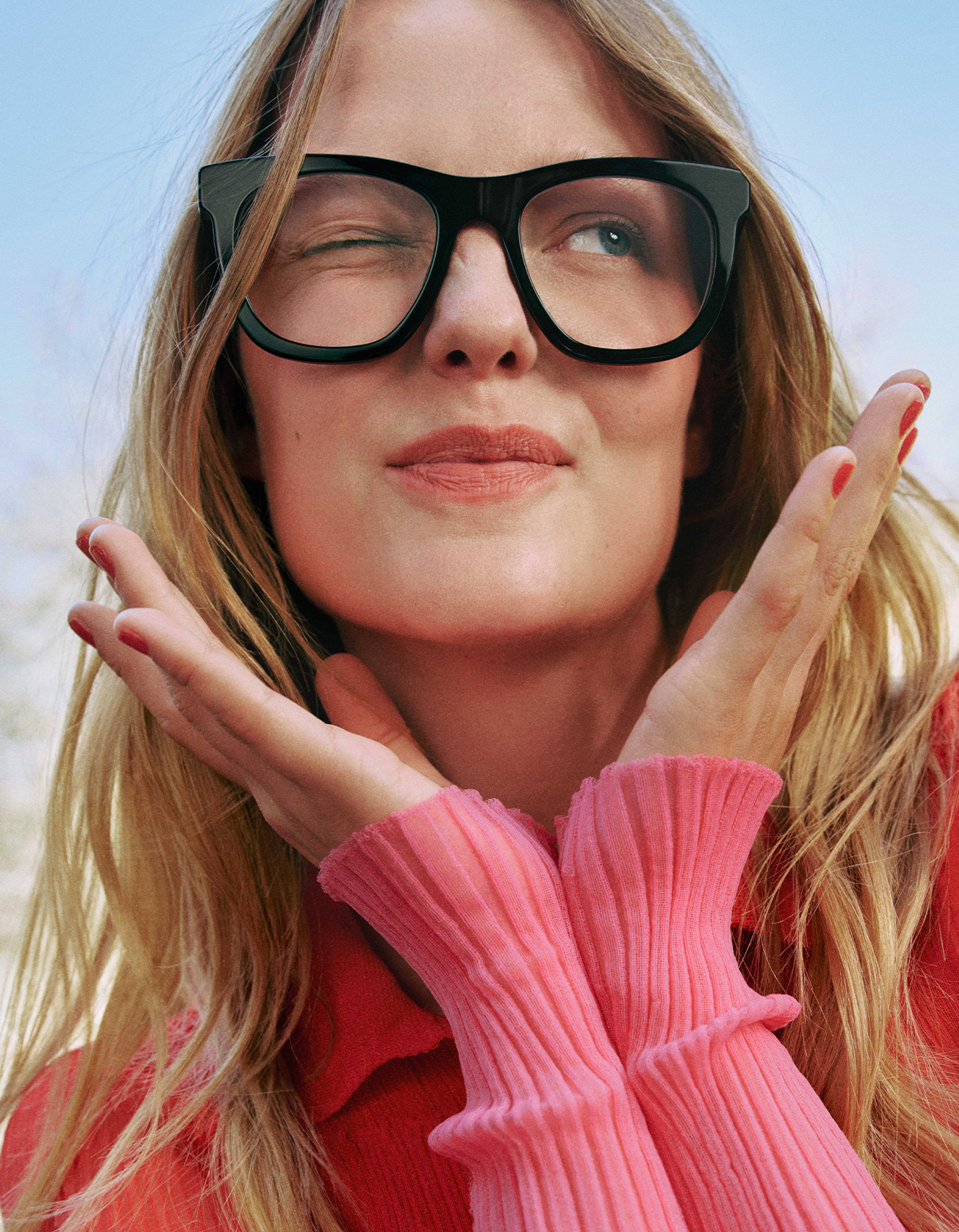 Una donna che fa l'occhiolino, indossa grandi occhiali neri e un maglione rosa e si copre il viso con entrambe le mani sullo sfondo di un cielo di un blu splendente.
