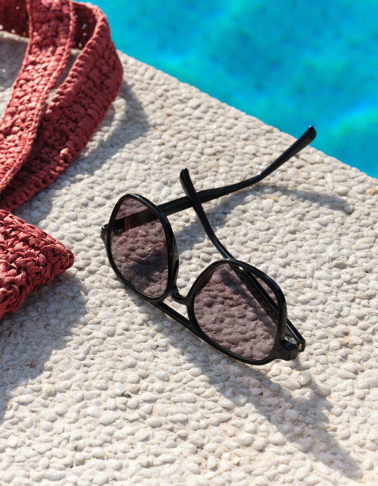 Un paio di occhiali da sole neri con lenti colorate giace su una superficie ruvida accanto a una piscina. A sinistra si intravede in parte una borsa rossa in tessuto.