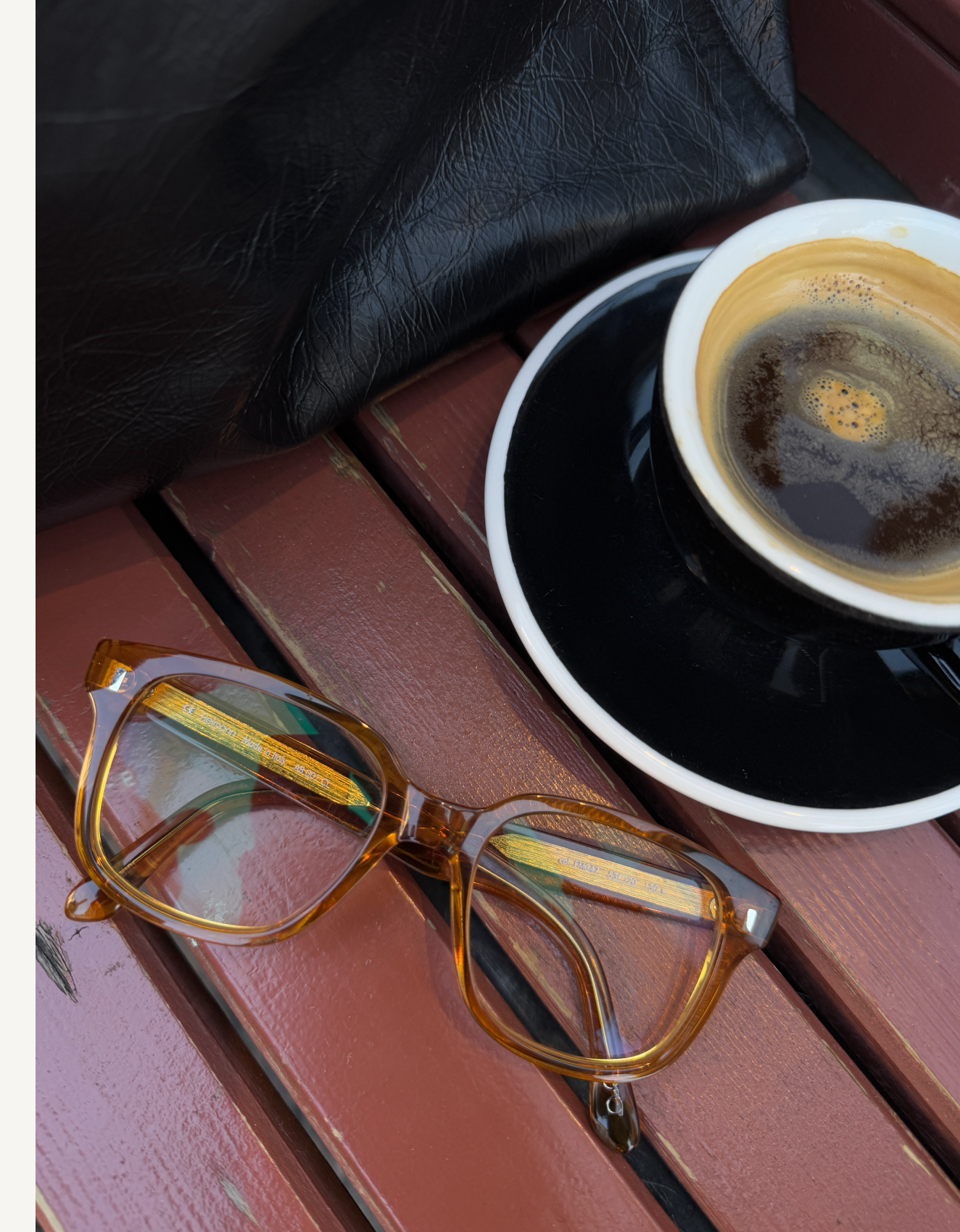 Occhiali con montatura marrone accanto a una tazza di caffè nero, posati su un tavolo di legno con una giacca di pelle nelle vicinanze.