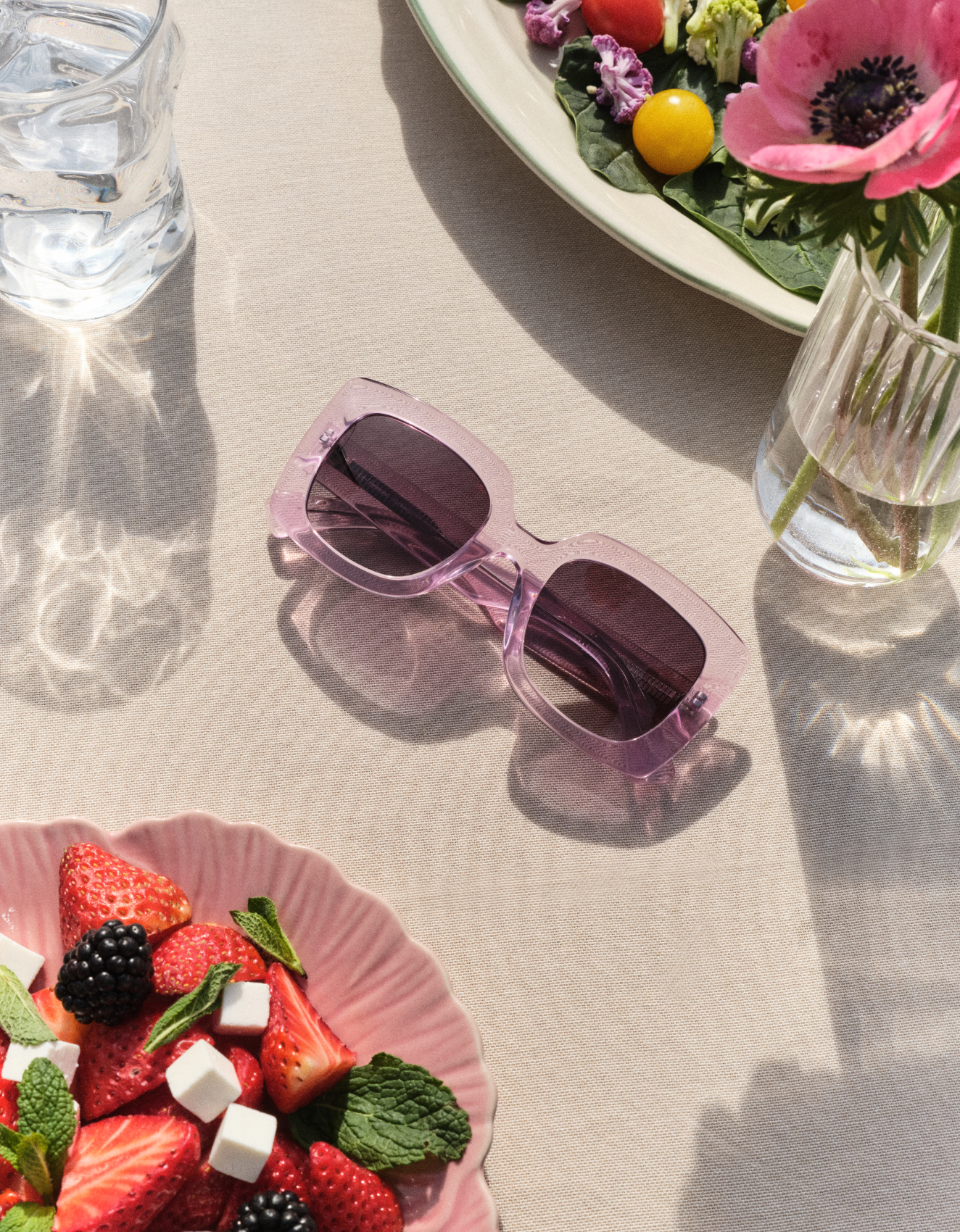 Różowe okulary przeciwsłoneczne na beżowym obrusie, otoczone talerzem z truskawkami, sałatą liściastą i szklanką wody.