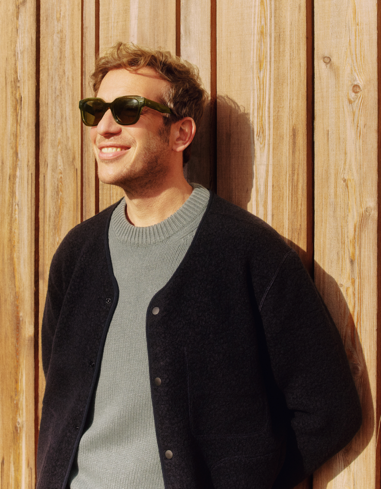 Ein Mann mit Sonnenbrille steht lächelnd vor einer Holzwand und genießt die Sonne. Er trägt einen grauen Pullover und eine schwarze Jacke.