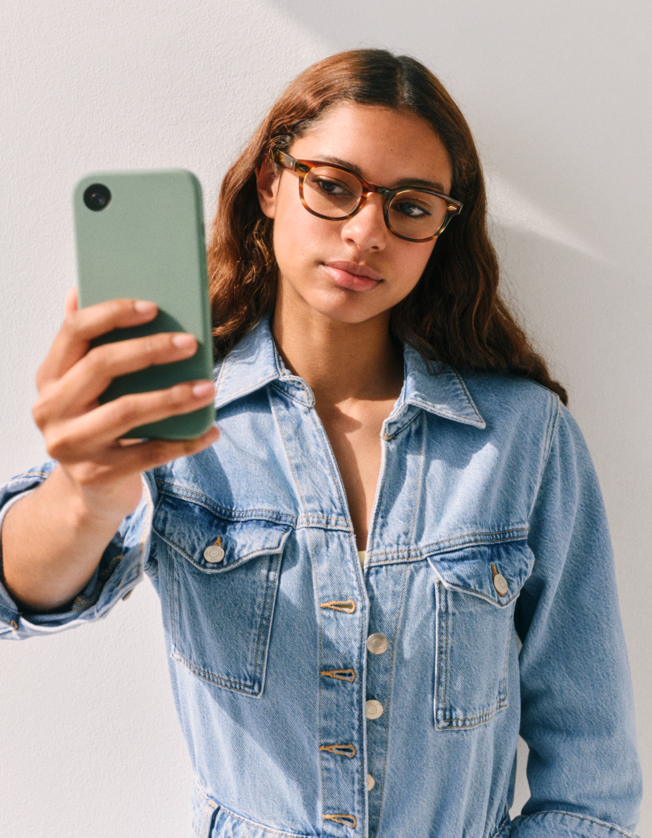Una persona con gli occhiali e una giacca di jeans che si scatta un selfie con uno smartphone verde su uno sfondo bianco.