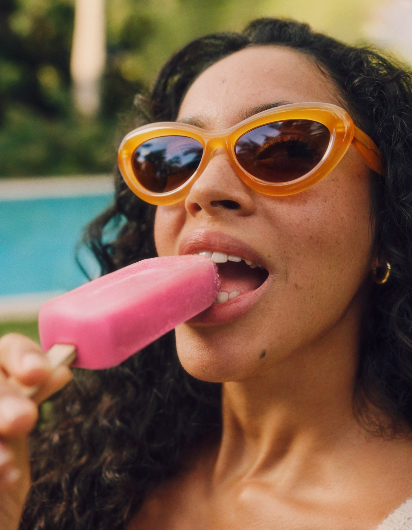 Frau mit orangefarbener Sonnenbrille, die draußen ein rosa Eis am Stiel genießt. Lockiges Haar, große Creolen, sonniger Hintergrund.