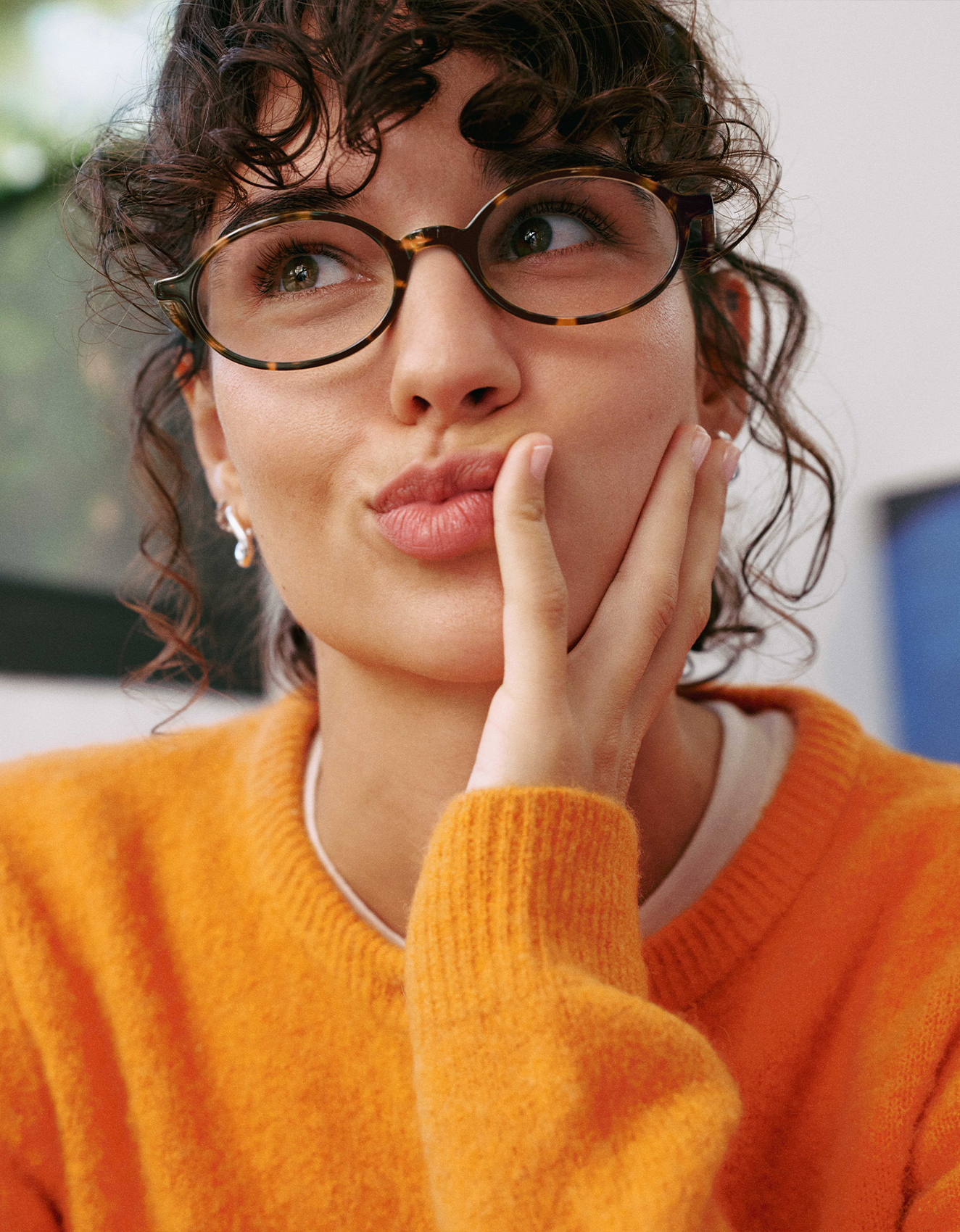 Una persona con i capelli ricci e gli occhiali, che indossa un maglione arancione, fa una smorfia e si tocca la guancia con una mano.