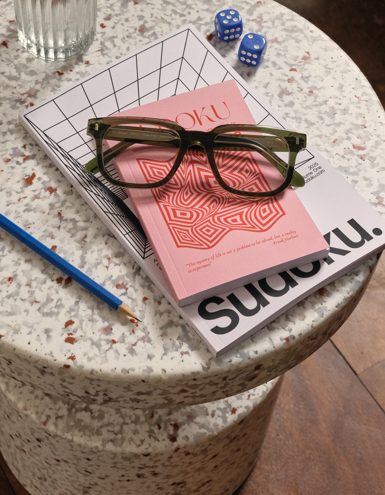 Eine Brille, Sudoku-Rätselhefte, ein Bleistift und Würfel auf einem gesprenkelten runden Tisch.