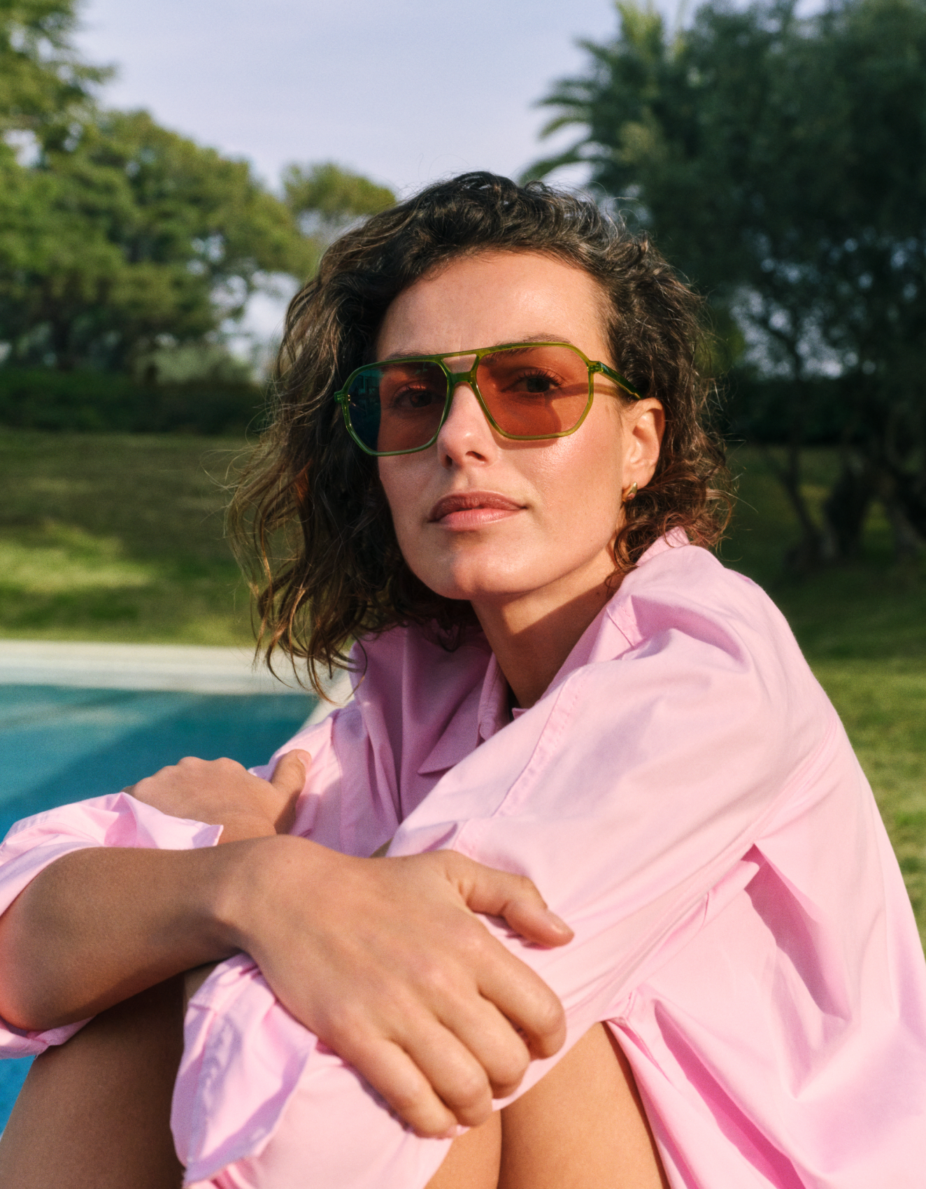 Una donna con una maglietta rosa e gli occhiali da sole è seduta a bordo piscina, con degli alberi sullo sfondo, e si gode la giornata di sole.