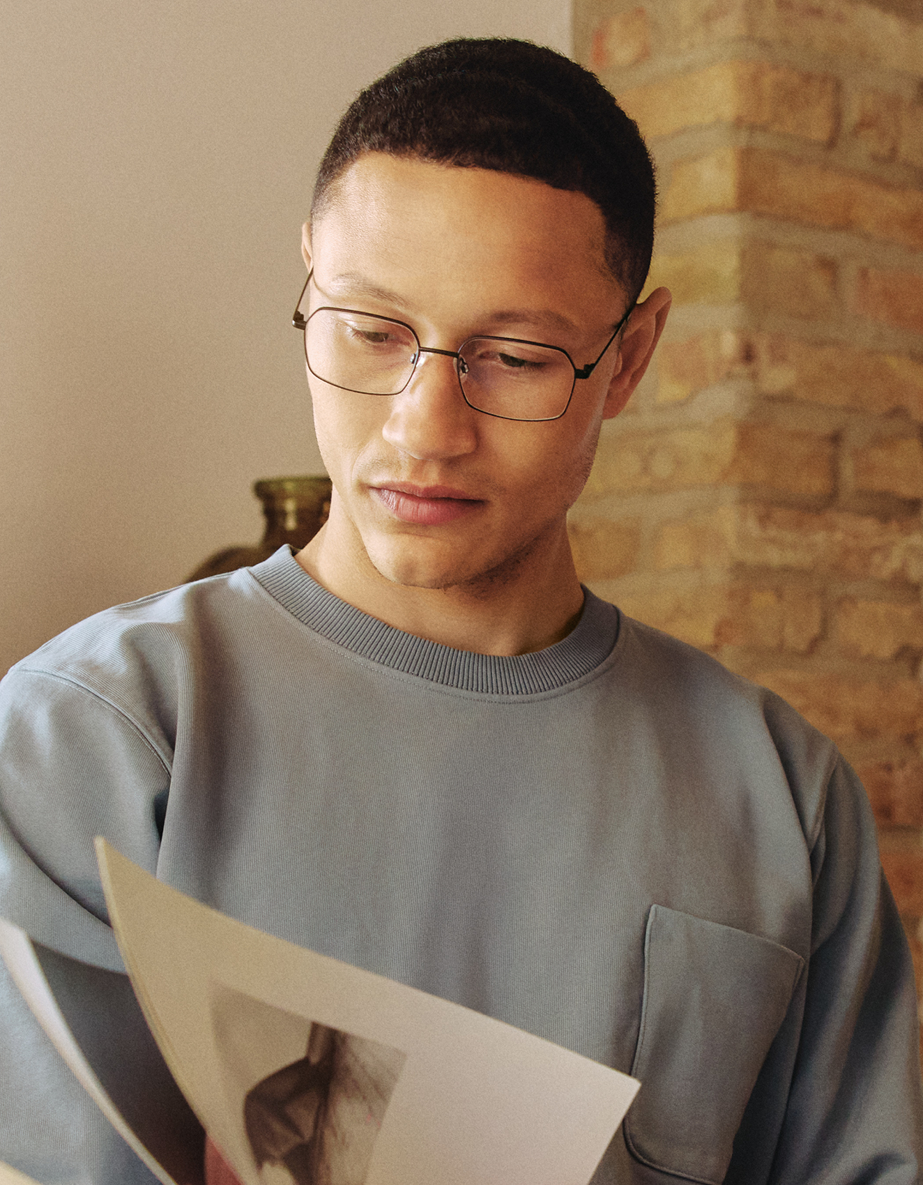Ein Mann mit Brille und blauem Pullover betrachtet eine Zeitschrift und steht drinnen neben einer Ziegelwand.